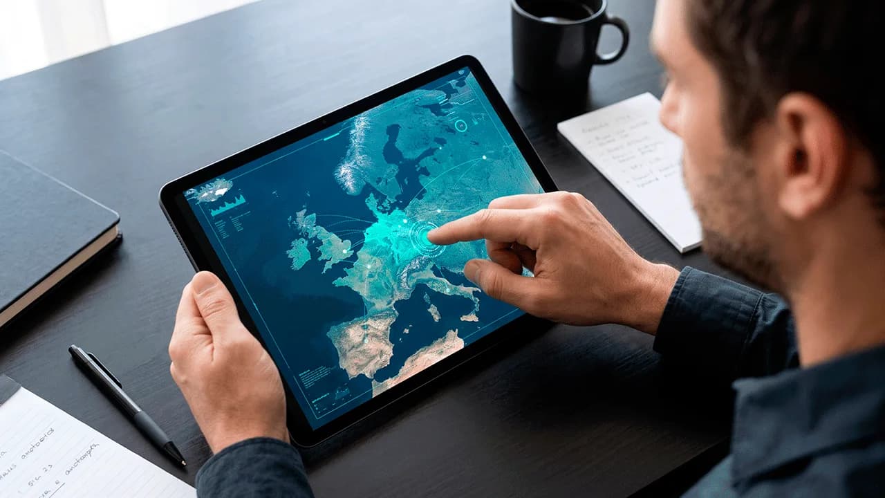 Homem sentado à mesa de madeira escura usando um tablet que exibe um mapa interativo da Europa em tons de azul.
