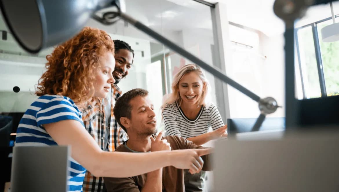 Quatro colegas de trabalho reunidos em frente a um computador, sorrindo e discutindo algo na tela em um ambiente de escritório moderno