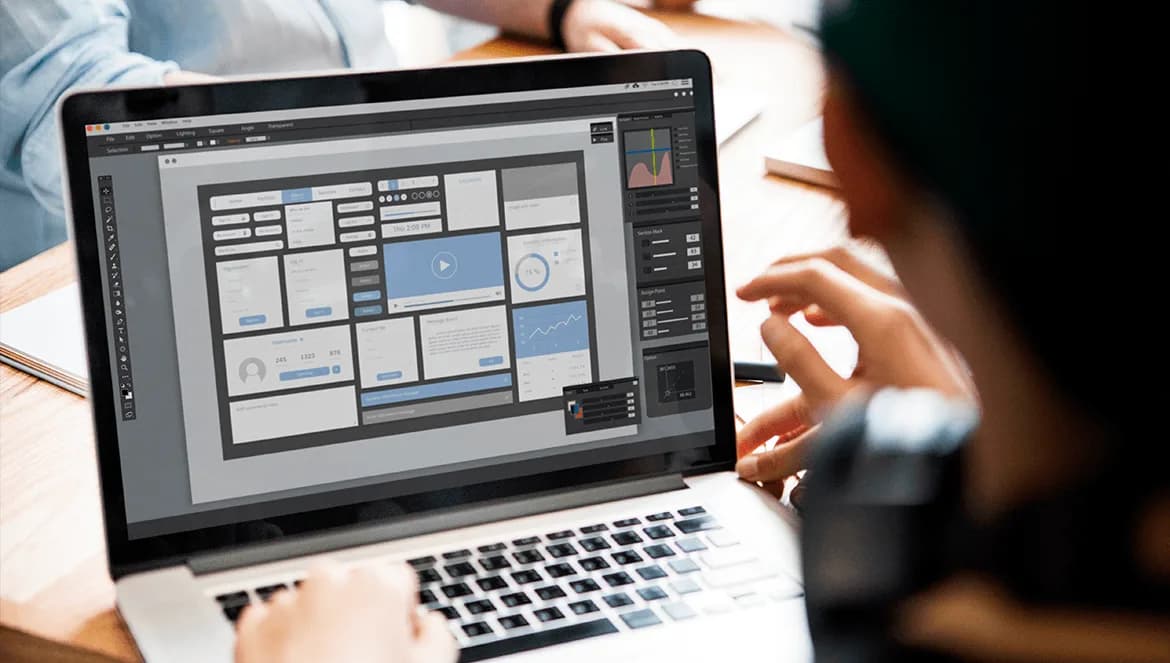 Visão por cima do ombro de uma pessoa trabalhando em um notebook. A tela do computador está em foco e exibe um software de design de interface (UI/UX), onde é possível ver o wireframe de um painel (dashboard) com diversos elementos estruturais, como blocos de layout, gráficos, áreas de perfil e um player de vídeo.