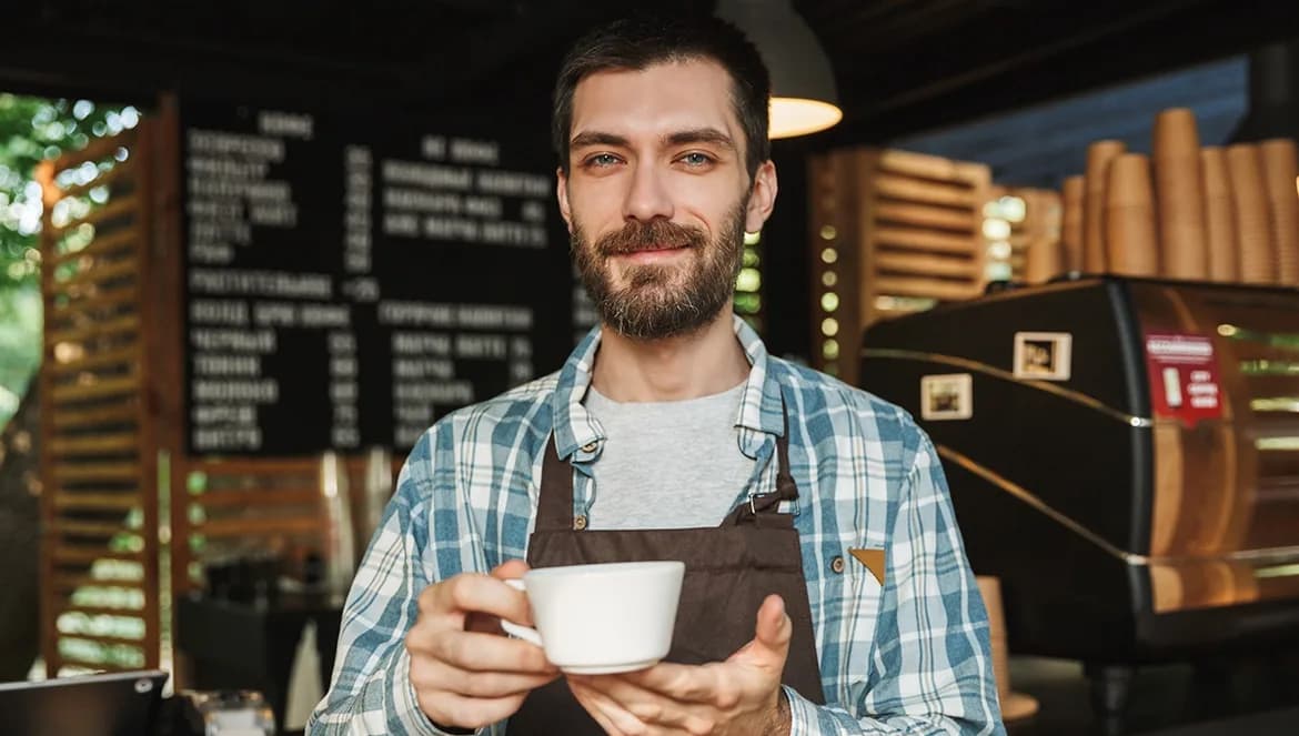 Um barista sorridente com barba e avental marrom segurando uma xícara de café branca com as duas mãos, oferecendo-a para a câmera. Ao fundo, o ambiente acolhedor de uma cafeteria. Tudo isso ilustrando um artigo sobre marketing digital.