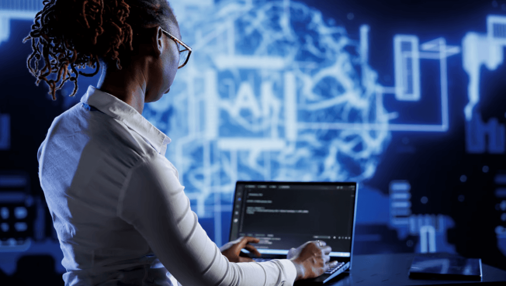Pessoa trabalhando em um laptop com códigos na tela, em um ambiente tecnológico iluminado por um grande painel azul que exibe ilustrações digitais de um cérebro e circuitos, sugerindo temas de IA e computação avançada.